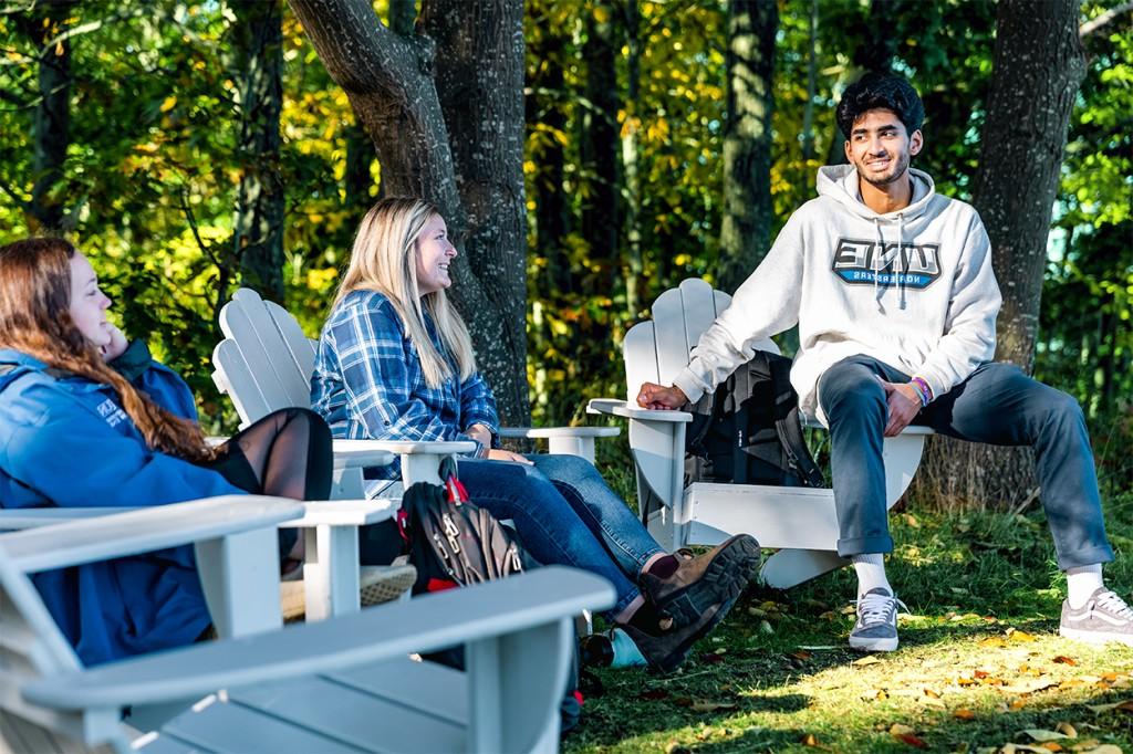 Three students sitting in adirondack chairs outside in front of the woods