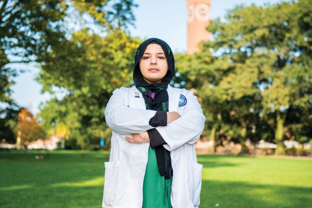 Fajar in her hijab and U N E C O M white coat with arms crossed, standing outside on a bright summer day