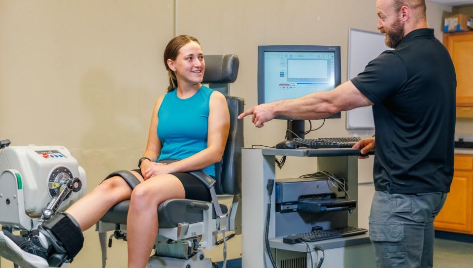 一名学生坐在一台测量肌肉力量的机器上，教授在显示器上向他们展示结果