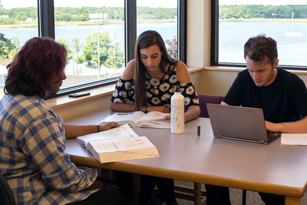 Three U N E students study together a table next to windows with a view of the ocean