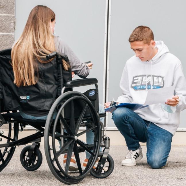 A student holding a clipboard kneels down next to another student in a wheelchair